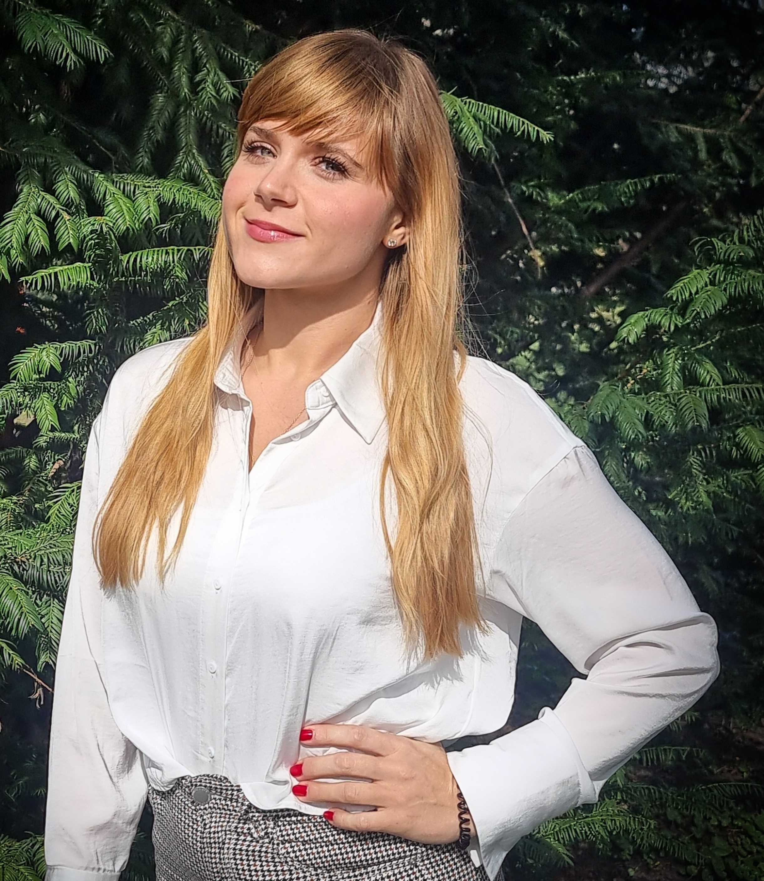 Lächelnde Frau mit weißer Bluse steht draußen im Grünen.